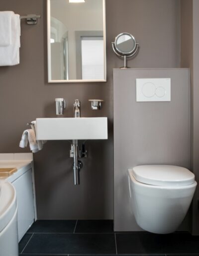 Links an der Wand befindet sich die Badewanne mit einem Wasserschutz aus Plexiglas. Rechts daneben das Lavabo mit anschliessendem WC. Über dem Lavabo hängt ein Spiegel.