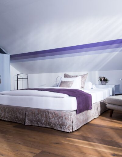 Ein Bild von einem Hotelzimmer. Links ist ein hellvioletter Einbauschrank. Rechts davon steht ein grosses Doppelbett mit Nachttischchen. Anschliessend ist ein grauer Sessel mit Fussteil und Stehlampe.