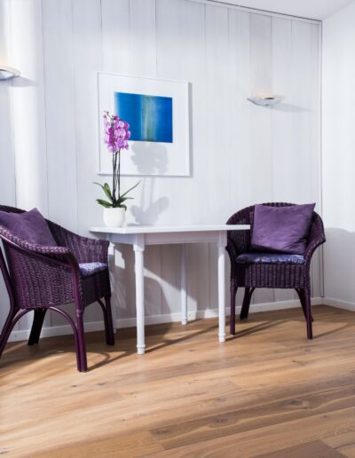 An einer grauen Bretterwand steht unter einem Bild ein weisser Tisch. Darauf steht eine Orchidee in einem weissen Topf. Auf beiden Seiten des Tisches steht jeweils ein Stuhl. An der rechten Wand hängt ein Spiegel und auf der linken Seite geht's auf den Balkon.